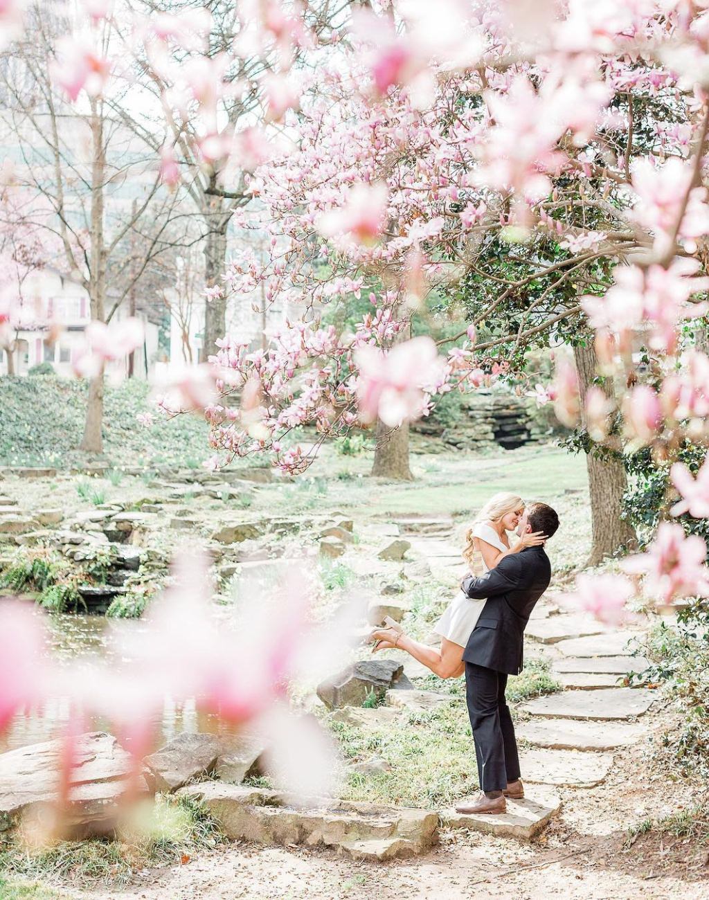 A guy picking up a girl and spinning her under spring tree blossoms. They are taking pictures for their engagement session