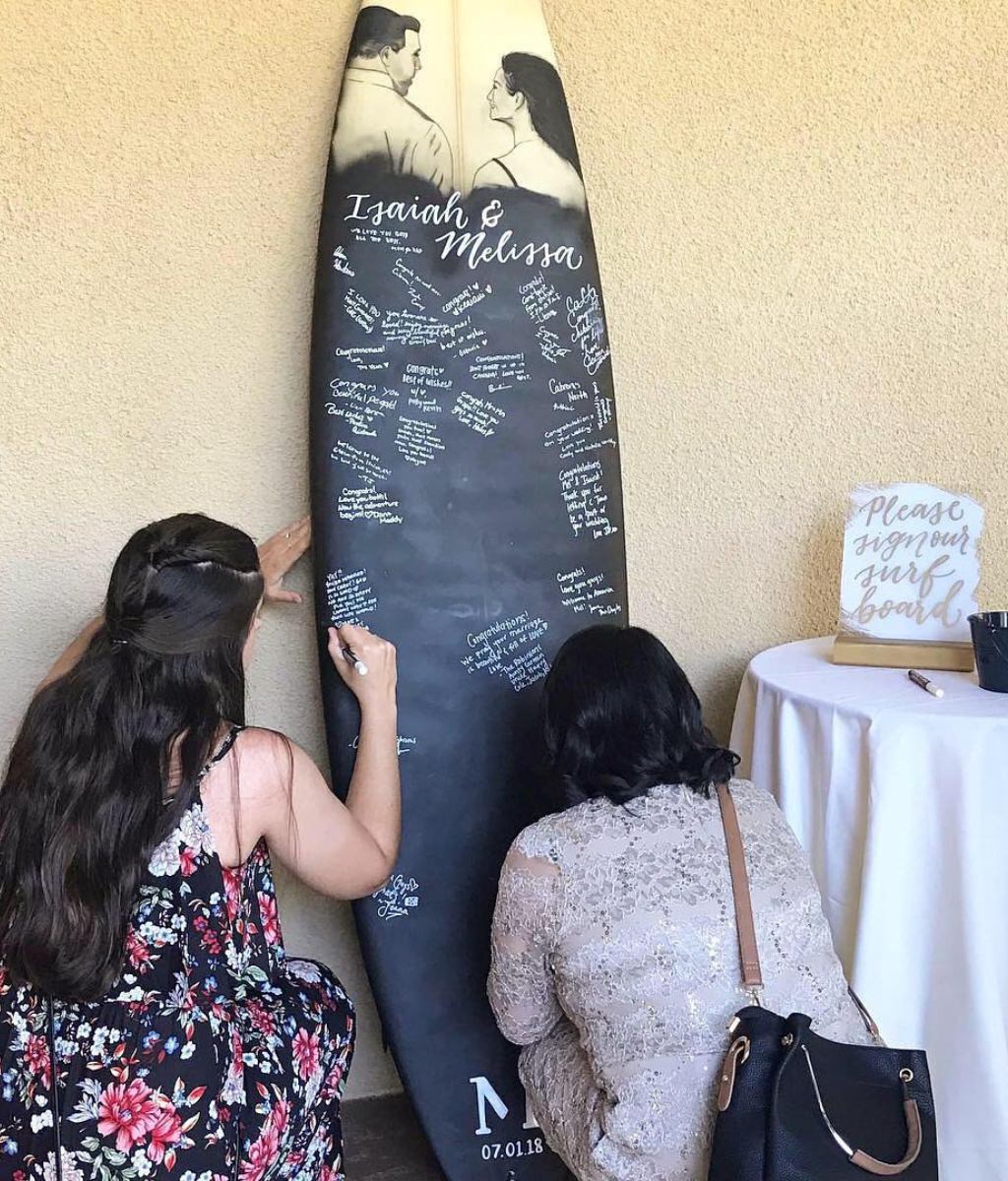 Wedding guests signing a full-sized surfboard that the wedding couple is using for their guest book. This is a fun and unique alternative ideas to the traditional guest book.